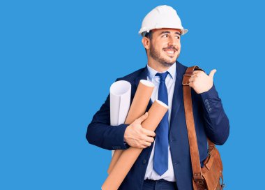 Mimar şapkalı ve deri çantalı genç İspanyol adam elinde planlar, başparmağıyla yan tarafa bakıyor ve ağzı açık gülümsüyor. 