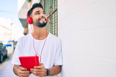 Genç Arap adam şehirde kulaklık ve dokunmatik ped kullanarak mutlu bir şekilde gülümsüyor..
