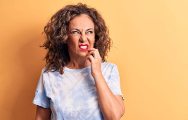 Orta yaşlı, güzel bir kadın, sarı arka planda dikilen günlük boyalı tişört giyiyor, şüpheye odaklanmış, çenesinde parmak olan ve kafasını kaldırıp,