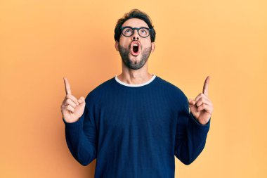 Gündelik giysiler ve gözlük takan genç İspanyol adam şaşırmış ve şaşırmış bir şekilde yukarı baktı ve parmaklarıyla işaret etti ve kollarını kaldırdı.. 