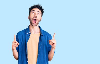 Gündelik giysiler giyen genç İspanyol adam şaşırmış ve şaşırmış bir şekilde yukarı bakıyor ve parmaklarıyla işaret ediyor ve kollarını kaldırıyor.. 