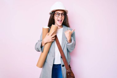 Şirin İspanyol çocuk mimar şapkası takmış elinde planlar, başparmağıyla yan tarafı işaret ediyor ağzı açık gülümsüyor. 