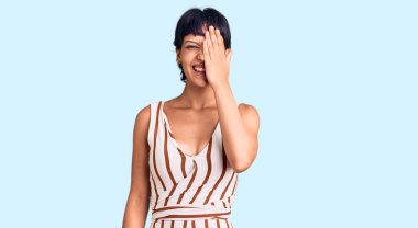 Kısa saçlı, kısa saçlı, yaz elbisesi giyen, bir gözünü eliyle kapatan, yüzünde kendine güvenen bir gülümseme ve şaşırtıcı bir duygu olan genç esmer kadın.. 