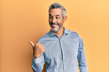 Orta yaşlı, gri saçlı, günlük kıyafetler giyen, gülümseyen, gülümseyen ve baş parmağıyla yüzünü gösteren bir adam.. 