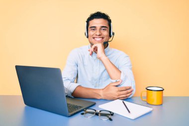 Young handsome hispanic man working at the office wearing operator headset smiling looking confident at the camera with crossed arms and hand on chin. thinking positive.  clipart