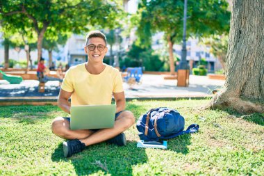 Üniversite kampüsünde çimlerin üzerinde otururken mutlu bir şekilde gülümseyen İspanyol öğrenci.