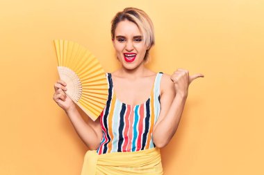 Mayo giymiş genç, sarışın bir kadın el ele tutuşmuş yelpaze parmağıyla yan tarafı işaret ediyor mutlu bir şekilde gülümsüyor. 