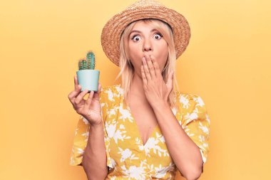 Sarı arka planda kaktüs saksısı tutan güzel sarışın kadın ağızlarını eliyle kapatıyor, şok geçiriyor ve hata yapmaktan korkuyor. Şaşırtıcı ifade.