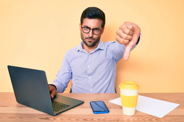Young hispanic man working at the office drinking a cup of coffee with angry face, negative sign showing dislike with thumbs down, rejection concept