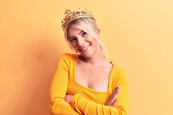 年轻美丽的金发女子戴着金色的公主王冠 披着孤零零的黄色背景 开心的脸上挂着微笑 交叉着双臂看着相机 积极的人 — 图库照片