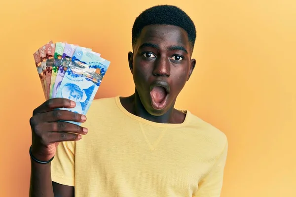 Young African American Man Holding Canadian Dollars Scared And Amazed young-african-american-man-holding-canadian-dollars-scared-and-amazed