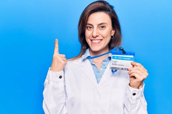 Young beautiful woman wearing doctor stethoscope holding id card smiling with an idea or question pointing finger with happy face, number one