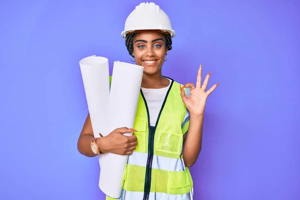 Young african american woman with braids wearing safety helmet holding blueprints doing ok sign with fingers, smiling friendly gesturing excellent symbol