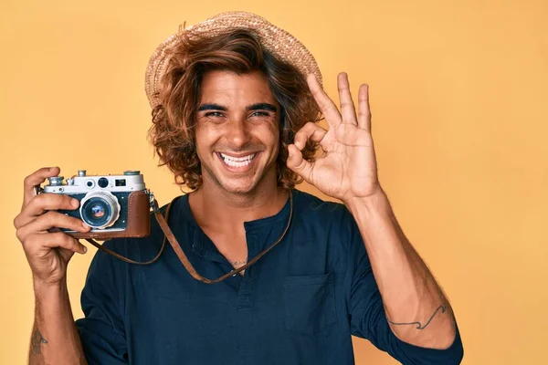 Young hispanic man wearing hat holding vintage camera doing ok sign with fingers, smiling friendly gesturing excellent symbol