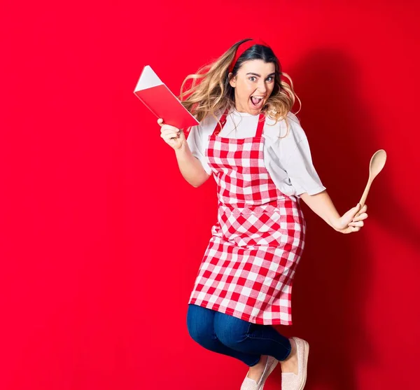 Young beautiful blonde cook woman wearing apron smiling happy. Jumping with smile on face holding recipe book and spoon over isolated red background