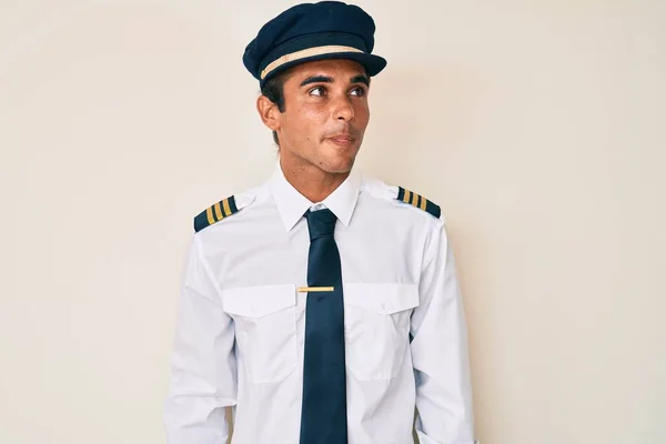 飛行機のパイロットの制服を着た若いヒスパニック系の男が横を見て笑って — ストック写真