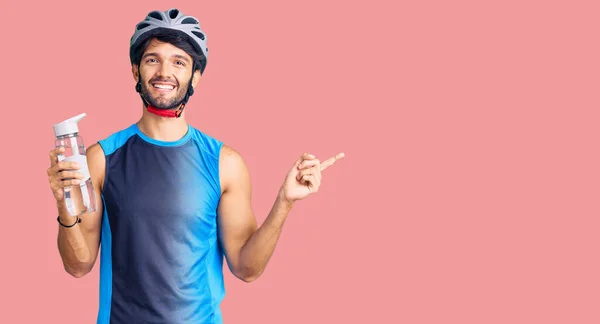 Handsome hispanic man wearing bike helmet and holding water bottle smiling happy pointing with hand and finger to the side