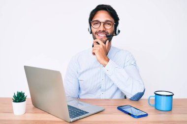 Ofiste çalışan yakışıklı İspanyol bir adam operatör kulaklığı takıyor kameraya kollarını kavuşturmuş ve çenesinde elle gülümsüyor. Olumlu düşünmek. 