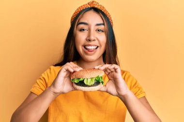 Lezzetli bir klasik burger yiyen genç Latin kadın yüzünde mutlu ve havalı bir gülümsemeyle gülümsüyor. diş gösterme. 