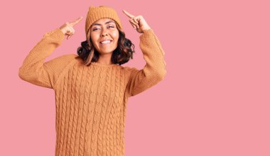 Genç, güzel, melez bir kadın yün kazak ve kış şapkası giyiyor. İki eliyle başını gösteren bir gülümseme. Harika bir fikir ya da düşünce, iyi bir hafıza. 