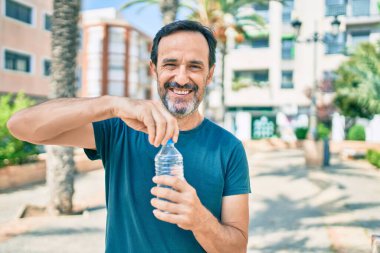 Orta yaşlı, sakallı, mutlu, açık havada su içen bir adam.