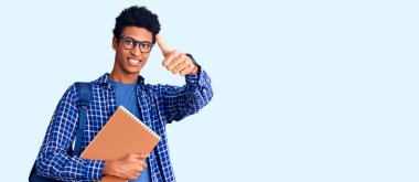 Afrika kökenli Amerikalı genç bir adam sırt çantası takıyor ve elinde kitap tutuyor. Elinde olumlu bir jest yapıyor. Başparmağıyla gülümsüyor ve başarı için seviniyor. kazanan jest. 