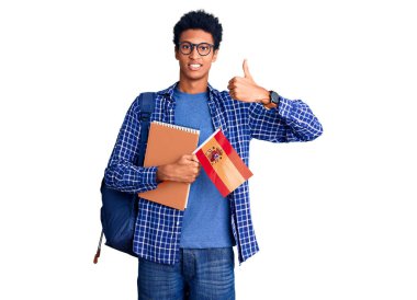 Afrika kökenli Amerikalı genç bir adam sırt çantasıyla İspanyol bayrağı taşıyor mutlu ve pozitif gülümsüyor, başparmağıyla mükemmel ve onay işareti yapıyor. 