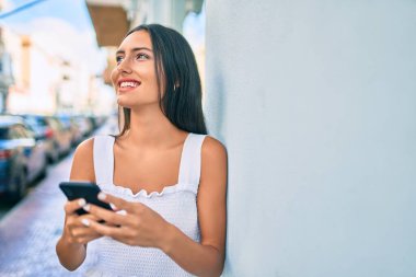 Genç Latin kız mutlu bir şekilde gülümsüyor. Duvara yaslanmış akıllı telefon kullanıyor..