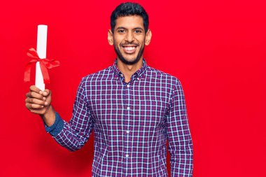 Diploma almış genç bir latin. Pozitif ve mutlu görünüyor. Dişlerini gösteren bir gülümsemeyle ayakta duruyor ve gülümsüyor. 