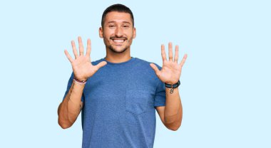 Dövmeleri olan yakışıklı bir adam. Günlük giysiler giyiyor. Kendini beğenmiş ve mutlu bir şekilde gülümserken on numarayı gösteriyor.. 