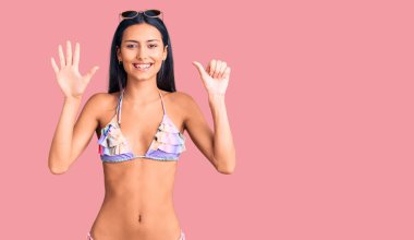 Bikini giyen, güneş gözlüklü genç ve güzel bir Latin kız. Altıncı parmaklarını gösterip gösterirken kendinden emin ve mutlu gülümsüyor.. 