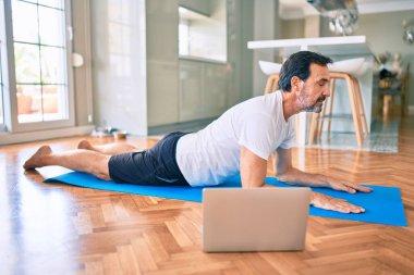 Orta yaşlı, sakallı ve esnek bir adam evde egzersiz yapıyor bilgisayardaki spor videolarına bakıyor.