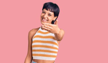 Kısa saçlı esmer bir kadın günlük kıyafetler giyiyor. Gülümsüyor, dostça el sıkışarak selamlıyor ve hoş karşılıyor. başarılı iş. 