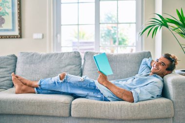 Genç İspanyol gülümseyen mutlu bir kitap okuyucu evdeki kanepede uzanıyor.