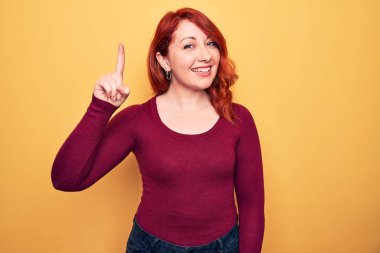 Genç, güzel, kızıl saçlı bir kadın sarı arka planda dikilmiş gülümsüyor bir fikir ya da soruyla mutlu bir yüzle işaret ediyor.