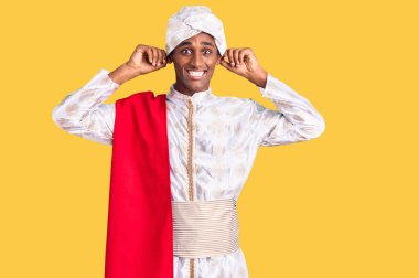 Geleneksel Sherwani sari giysileri giyen Afrikalı yakışıklı adam parmaklarıyla kulaklarını çekerek gülümsüyor, komik bir jest. deneme sorunu 