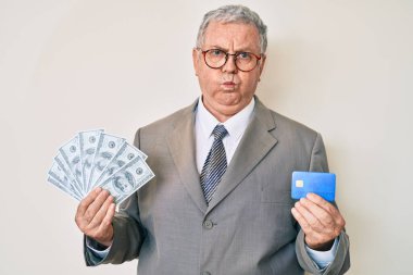 Üstün gri saçlı, takım elbiseli, kredi kartı ve komik suratlı, şişirilmiş yanakları olan bir adam. Ağzı havayla şişer, havayı yakalar.. 