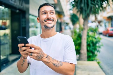 Şehrin caddesinde akıllı telefon kullanan, mutlu gülümseyen genç İspanyol adam..