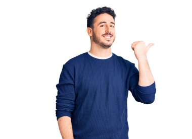 Gündelik kıyafetler giyen, gülümseyen, gülümseyen ve başparmağıyla yan tarafı gösteren İspanyol genç adam.. 