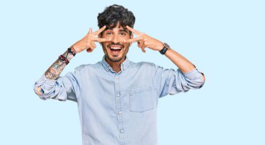 Gündelik kıyafetler giyen genç İspanyol adam barış sembolü yapıyor. Yüzünde parmaklar, gülümseyerek zaferi gösteriyor. 