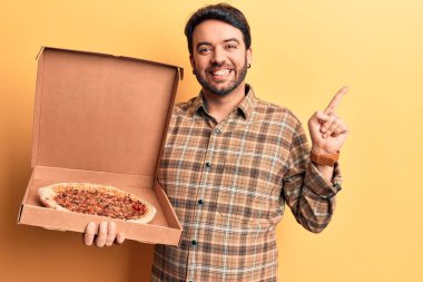 İspanyol asıllı genç adam pizza kutusunu tutarken gülümsüyor mutlu bir şekilde elini ve parmağını yana doğru gösteriyor. 