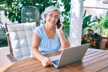 Gri saçlı, gülümseyen, mutlu, rahatlatıcı orta yaşlı bir kadın terasta oturuyor ve evden çalışıyor.