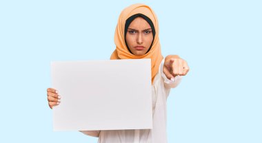 Genç esmer Arap kadın geleneksel İslami başörtüsü takıyor. Elinde kamerayı işaret eden bir pankart var. Kendine güveni tam bir jest. 