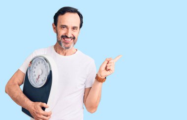 Orta yaşlı yakışıklı bir adam tartı makinesini tutuyor gülümsüyor mutlu bir şekilde elini ve parmağını yana doğru gösteriyor. 