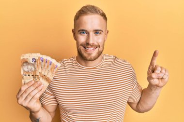 Güney Afrika 'da elinde 20 rand banknot tutan genç beyaz bir adam bir fikir veya soruyla gülümsüyor ve mutlu bir yüzle bir numarayı işaret ediyor. 