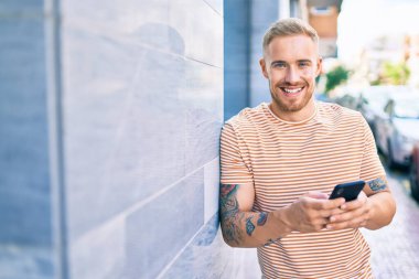 Genç İrlandalı mutlu bir şekilde gülümsüyor. Şehrin duvarına yaslanmış akıllı telefon kullanıyor..