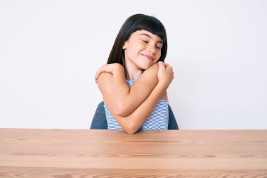 Çakma elbiseli genç bir kız masanın üzerinde oturmuş kendine sarılıyor mutlu ve pozitif, kendinden emin gülümsüyor. Sevgi ve özgüven. 