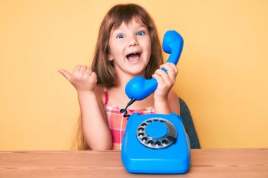 Masada oturan uzun saçlı beyaz bir kız. Klasik telefon kullanıyor. Başparmağıyla yan tarafı işaret ediyor. 