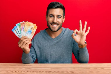 Yakışıklı İspanyol adam elinde İsviçre frangı banknotları tutuyor parmaklarıyla imza atıyor, gülümseyerek mükemmel bir sembol oluşturuyor. 