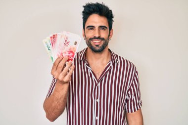 Sakallı yakışıklı İspanyol adam İsrail şekelleri tutuyor. Pozitif ve mutlu görünüyor. Kendine güvenen bir gülümsemeyle dişlerini gösteriyor. 